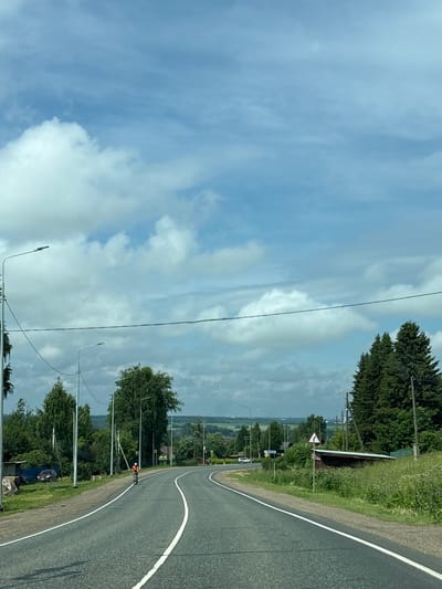 The image depicts a rural road curving to the right in Kstinin, Russia. The sky is partly cloudy with large cumulus clouds against a light blue background. Along the left side of the road, there are several tall streetlights and trees. A person on a bicycle, wearing a bright orange shirt and helmet, is cycling on the road ahead. Further in the distance, there are more trees, buildings, and rolling hills. On the right side of the road, there is a small shed with a triangular road sign indicating a sharp bend to the right. The road itself is asphalt with white lane markings. The overall scene suggests a sunny day in a countryside setting.