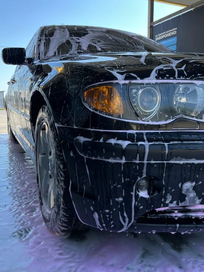 This is a close-up, low-angle shot of a black car being washed at a car wash in Velyki Hai, Ukraine. The car is covered in pink and white soap suds, which are dripping down the hood and bumper. The sky is a clear blue. A sign in the background, written in Ukrainian, reads "ON THE TERRITORY OF THE CAR WASH: FORBIDDEN:". The image focuses on the front passenger side of the vehicle, showcasing the headlight and wheel. The ground is covered in foamy water.