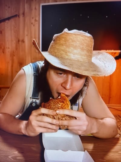 This media file shows a person wearing a straw hat and denim overalls, eating a sandwich. The person is leaning back, with their arm extended as they take a bite of the sandwich, which is wrapped in brown paper. The background is a wooden interior, possibly a cabin or shed, with a dark rectangular shape that could be a window or a framed picture. The overall lighting suggests an indoor setting. The image is rotated, making it appear as if the person is lying down or in an unusual orientation. The person's face is partially obscured by the hat and the sandwich, but they appear to be focused on eating. No text is visible in the image.