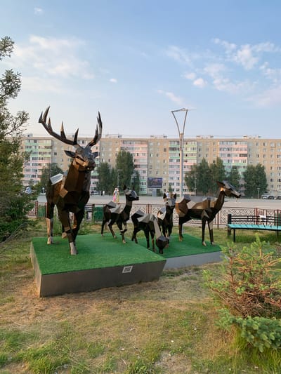 This image showcases a sculpture of a stag and a group of deer, crafted from faceted metal. The stag sculpture, prominently displayed in the foreground, is detailed with large antlers. Behind it, several smaller deer sculptures are positioned in a line, appearing to be in motion. The sculptures are set against a backdrop of a residential area, featuring tall, multi-story apartment buildings with rows of windows. Trees and greenery are visible around the sculptures and in front of the buildings. The scene is outdoors, under a bright sky with scattered clouds, suggesting daytime. A lamppost stands tall in the background. There is a QR code visible on a black base in front of the sculptures, which is an object that could provide more information about the artwork or its location. The overall impression is that of an urban art installation in a community setting.