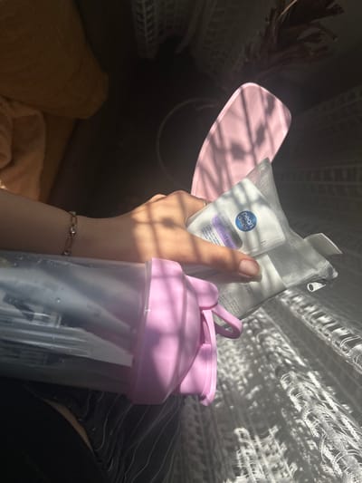 A close-up shot shows a person's hand holding a small, zippered, mesh pouch containing personal care items. The pouch is white with a metallic sheen. Visible on the pouch are the words "Pearl Drops" and "Whitening Smile." To the left of the pouch is a clear plastic water bottle with a pink lid and strap. The person is wearing a bracelet on their wrist. The background suggests an indoor setting with dappled sunlight creating shadows on white, textured fabric, possibly curtains or bedding. The image is taken from a slightly elevated angle. The location is not discernible from the image.