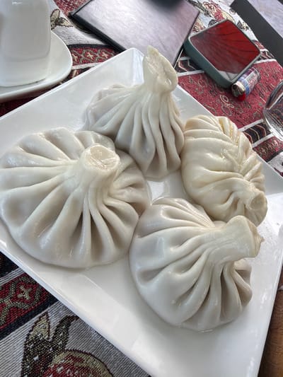 This is a close-up, overhead shot of a white ceramic plate containing four khinkali, a traditional Georgian dumpling, served on a table with a red and white patterned tablecloth. The khinkali are plump and appear to be freshly steamed, with a visible broth pooling at the bottom of each one. The dough is a pale, almost translucent white, and the pleated tops are gathered to a point. To the left of the plate, a white ceramic teacup and saucer are partially visible. In the background, a dark, rectangular object, possibly a menu or a tablet, rests on the tablecloth, next to a red object that resembles a lighter. The lighting is bright, suggesting it's daytime, and the overall scene is indoors. The image was taken in Kaluđerac, Montenegro.