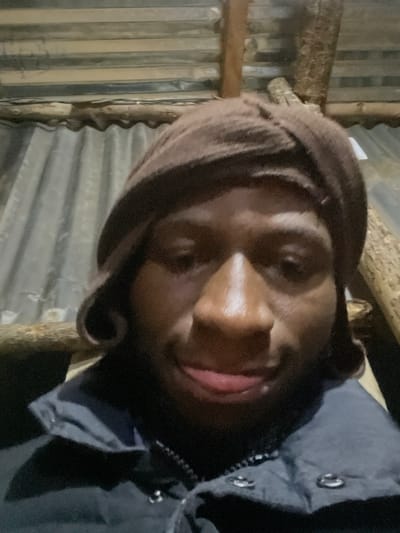 The image is a close-up, selfie-style photograph of a young Black male wearing a brown beanie and a dark jacket. He is looking directly at the camera with a slight smile and his tongue slightly protruding. The background appears to be an indoor setting with a corrugated metal roof and wooden beams. The lighting is dim and appears to be artificial. The location is identified as Agwala, Nigeria.