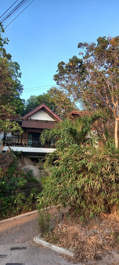 The image shows a building with a red-trimmed gable roof in Baan Wok Tum, Thailand. The building is surrounded by trees, including bamboo, and is situated on a rocky outcrop. The sky is clear blue. A concrete road is visible in the foreground. Power lines are visible in the top left.