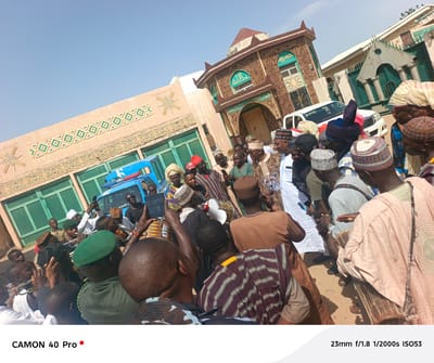 A low-angle shot depicts a gathering of people, mostly men, in front of a building with distinct architectural designs. Some people are in the foreground, facing away from the camera. Many are playing drums and musical instruments, and some are recording with their phones. The building is in Wase, Nigeria. It features geometrical designs in brown and blue-green and has an Islamic inscription in Arabic on the building's facade. A blue police car and a white car can be seen behind the crowd. The sky is clear and blue, indicating a sunny day.