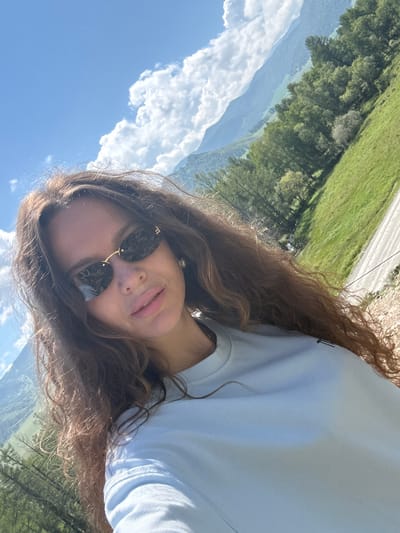 A medium shot shows a young woman with long, wavy brown hair, wearing dark sunglasses and a light blue t-shirt, posing for a selfie outdoors. She is looking at the camera with a slight smile. Her hair is partially covering her face and shoulders. Behind her, a vast landscape unfolds under a clear blue sky with fluffy white clouds. Rolling green hills covered with trees dominate the background, and mountains are visible in the distance. The terrain appears to be natural and undeveloped, suggesting a rural or mountainous setting. The image does not provide specific details that can identify the location as Bichiktu-Boom, Russia.