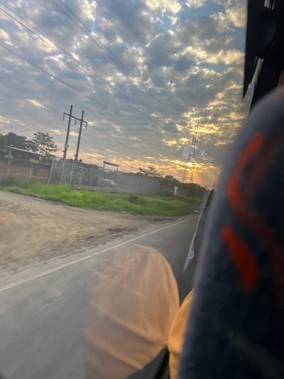 The image captures an exterior scene from the interior of a vehicle moving along a paved road in Milagro, Ecuador, during either sunrise or sunset. The sky is predominantly filled with a dense layer of altocumulus clouds, which are illuminated with golden light near the low-lying sun on the right horizon. Multiple overhead power lines traverse the upper portion of the frame.

On the left side of the scene, a wooden utility pole with cross-arms supports several wires. Below it, a low brick or concrete wall topped with chain-link fencing runs parallel to the road. Within this fenced area, structures and a white object resembling a partial fuselage or large industrial equipment are visible. The immediate ground beside the paved road consists of an unpaved dirt shoulder and a grassy verge.

Towards the right, a tall metal lattice transmission tower is silhouetted directly against the intense golden light of the sun. Distant trees line the horizon. Inside the vehicle, a dark blue fabric object with orange patterns, consistent with a seat or interior panel, partially obscures the right side of the view. A faint, light-colored reflection is visible on the window glass in the lower-middle portion of the image.
