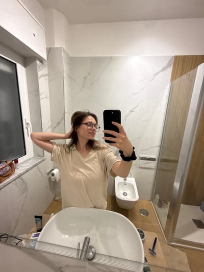 This image depicts a residential bathroom in Scafati, Italy, captured via a mirror selfie.

**Person:** A female, estimated to be a young adult, is visible in the mirror's reflection. She has medium-brown, shoulder-length hair and wears dark-framed glasses. She is dressed in a light beige or cream-colored V-neck short-sleeved top. Her left arm is raised, with her hand positioned near the back of her head, lightly touching her hair. Her right hand holds a black smartphone, positioned to capture the mirror reflection. A black scrunchie is worn on her right wrist. She exhibits a slight smile.

**Environment:** The bathroom features white walls with grey marble-like veining. The floor and a built-in counter surface under the sink are composed of light-colored wood or wood-look material. The ceiling is white, and the room is brightly illuminated by artificial light sources. A white-framed window is visible on the far left, offering a view of darkness beyond, suggesting nighttime. An overhead white cabinet is mounted above the window.

**Objects and Fixtures:**
*   **Foreground (reflected):** A white ceramic vessel sink with a chrome-finished faucet is prominent. On the wooden surface to the right of the sink, a black tube (possibly a cosmetic product), a small black circular container, and a blue disposable razor are visible.
*   **Midground:** A white wall-mounted toilet paper holder with a roll of white toilet paper is present on the left. A white ceramic bidet with a chrome faucet is situated to the right of the toilet paper holder.
*   **Background Right:** A glass shower enclosure with chrome framing is visible. Inside the shower, on a shelf, several plastic bottles (likely toiletries) are arranged. A section of the wall adjacent to the shower features vertical light-brown wooden slats.
*   **Background Left (on windowsill):** A brown leather-look pouch or small bag and a red-capped bottle are resting on the white windowsill.

**Actions and Interactions:** The female subject is actively taking a mirror selfie with her smartphone, posing with one hand in her hair, indicating a conscious photographic capture of herself within the bathroom setting.