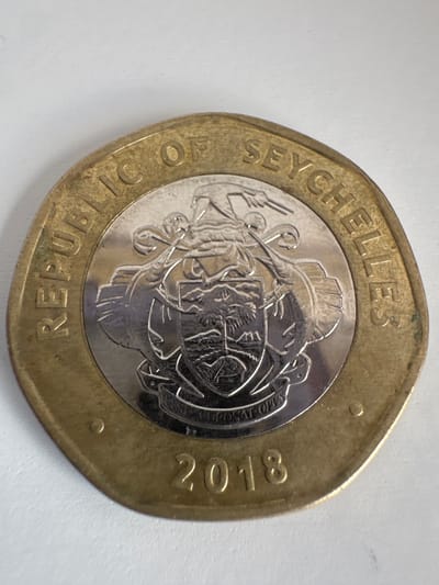 This close-up photograph captures the obverse side of a bimetallic coin from the Republic of Seychelles, dated 2018. The coin features an outer brass-toned ring and an inner silver-toned disc.
The outer ring bears the inscription "REPUBLIC OF SEYCHELLES" in capital letters, curving along the top and bottom edges. The year "2018" is prominently displayed on the right side of this outer ring.
The central inner disc showcases the detailed Coat of Arms of Seychelles. The emblem consists of a shield depicting a giant tortoise on green ground, a coco de mer palm tree, and a ship sailing on the ocean. This shield is supported on either side by two sailfish. Above the shield, a blue and white wreath with a red ribbon is topped by a white-tailed tropicbird in flight. A ribbon below the shield contains the national motto, with parts of "CORONAT OPUS" visible.
The coin exhibits some signs of wear and tarnishing. The scene is a static object shot against a plain white background, with no activity or event taking place. The photograph was taken in Subotica, Serbia.