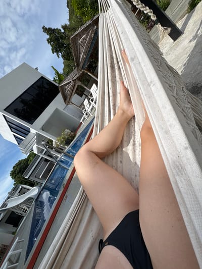 The image shows a person relaxing in a hammock outdoors. The person is wearing a black bikini and their legs are visible, with some water droplets on their skin, suggesting they may have recently been swimming or are by a pool. The hammock is made of white rope and fabric.

In the background, a resort-like setting is visible. There is a swimming pool with blue tiling and a metal ladder. Several white lounge chairs are placed around the pool area. A modern white building with large dark-tinted windows is also present, as are thatched-roof structures that could be gazebos or cabanas. Lush green trees and tropical foliage surround the area, indicating a warm climate. The sky is blue with some scattered white clouds. The sun appears to be shining, casting shadows on the ground, suggesting it is daytime. The overall scene evokes a sense of relaxation and vacation in a tropical location. The image is rotated 90 degrees counter-clockwise.
