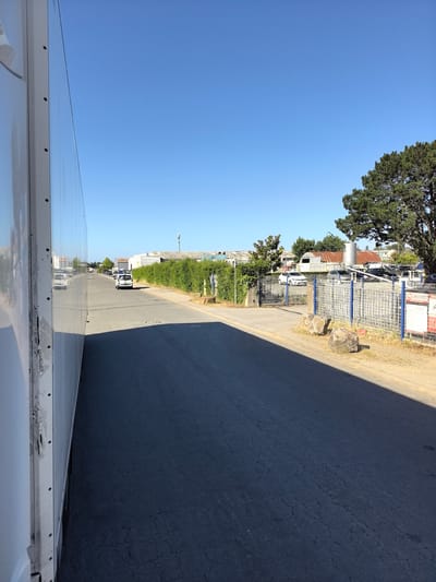 This image captures a street scene in Saint-Herblain, France, on a clear day. The viewpoint appears to be from within or adjacent to a large white vehicle, possibly a trailer or truck, occupying the left side of the frame. The vehicle's reflective surface dominates the foreground on the left. The road, surfaced with dark asphalt showing cracks, extends into the distance, leading towards a line of industrial buildings and a dense green hedge. On the right side of the road, a blue and silver metal fence encloses a lot with parked cars and some large rocks in front of it. A mature evergreen tree with dark green foliage stands tall behind the fence. A white utility vehicle or small bus is visible driving down the road in the middle distance, and other vehicles are parked in the lot behind the fence. The sky is a clear, bright blue.