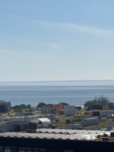 The image captures a wide-angle view of a coastal area, likely part of Loures, Portugal, characterized by a vast expanse of calm blue water meeting a hazy horizon. In the foreground, a large, dark building with a green roof covered in solar panels is visible. Behind it, a cluster of modular structures, shipping containers, and temporary event tents are spread across a grassy area. These structures appear to be set up for an event, with some adorned with large, colorful banners. Some construction or setup activities are ongoing, with a few individuals visible in the distance working around the temporary buildings and equipment. The overall scene suggests a daytime setting, with clear, bright skies and a generally tranquil atmosphere despite the presence of infrastructure for an event.