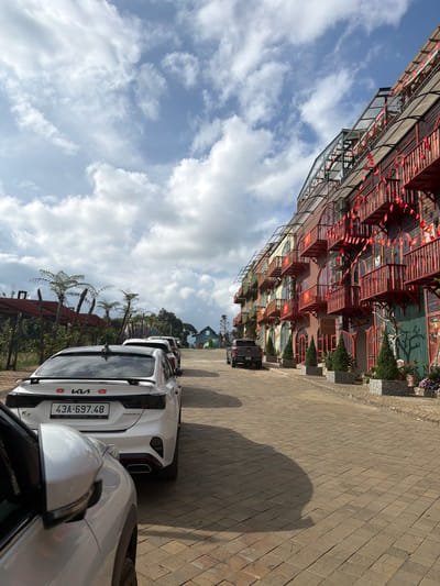 This outdoor image, taken in Măng Đen, Vietnam, features a distinctive multi-story building and several parked cars under a bright, partly cloudy sky. The building boasts unique and intricate architecture, characterized by numerous red-orange balconies and railings, glass elements, and a complex, layered design. Small red and yellow Vietnamese flags are strung decoratively along the building's facade and balconies, while planters with greenery and flowers add to its visual appeal.

A paved road or parking area occupies the foreground and mid-ground, where several cars are parked. The closest vehicle is a white Kia, with "KIA" and "PREMIUM" visible on its rear. Its license plate clearly displays "43A-697.48," along with smaller text "THACO KIA MOTORS" and a small Vietnamese flag symbol. Other cars, including a dark SUV, are parked further down the paved area. The ground is paved with rectangular blocks, and the presence of clear shadows indicates bright daylight. Palm-like trees or ferns are visible in the landscape around the building. The sky is a clear blue with scattered white clouds, suggesting pleasant weather. The scene portrays a unique architectural setting, likely a hotel or resort.