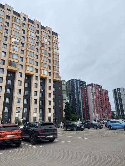 The image shows a parking lot in front of a row of modern apartment buildings, identified as being in Ligachevo, Russia. The sky is overcast. In the foreground, several cars are parked on a paved surface marked with white lines. The cars visible include an orange SUV on the left, a black luxury SUV in the center, and a dark blue SUV to its right. Behind these cars, a blue sedan is parked further to the right. In the background, a series of tall apartment buildings are visible. The building closest to the left is primarily light beige with dark grey sections and numerous balconies with orange trim. Further to the right, a collection of taller buildings with distinctive color schemes, including dark blue, black, red, and grey, are present. Some of these buildings also feature multiple balconies and windows. A few trees are visible between the buildings. There are no people clearly visible in the image.