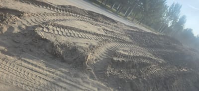 The image depicts a section of raw earth, likely a construction site or a path, characterized by prominent tire tracks from heavy machinery pressed into the sandy, rocky soil. The ground is uneven, with mounds and depressions indicating recent excavation or grading. In the background, a paved road is visible, bordered by a yellow line, with a row of trees lining the opposite side of the road. The trees are tall and slender, suggesting a park or natural area. The sky is bright and hazy, indicating daytime with possible atmospheric dust or light pollution. The overall scene suggests an outdoor environment undergoing development or maintenance, with no people or specific buildings identifiable within the frame. The location is identified as the city of Mramornoye, Kyrgyzstan.