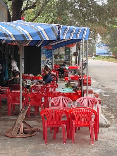 The image depicts an outdoor food service establishment situated on a paved area adjacent to a road in Thuận An Ward, Vietnam. A large blue and white striped umbrella or awning dominates the foreground, providing overhead cover. Beneath it, numerous identical red plastic chairs are arranged in rows and around small tables. Approximately two dozen chairs are visible, with many unoccupied.

Two individuals are present under the awning. On the left, a person wearing a dark top is seated with their back partially to the viewer, at a table displaying clear plastic containers. To their right, a second individual, dressed in a denim-like top and with hair tied back, is seated, actively engaged in handling items within a red plastic tray on a table in front of them. Surrounding these individuals are various red plastic tubs, baskets, and a larger light blue tub, containing what appears to be seafood or produce, placed on makeshift stands and directly on the ground.

Behind the establishment, a dark building facade features a large "Huda" beverage advertisement. A black motorcycle is parked to the left of the seated individuals. To the right of the outdoor setup, a paved road stretches into the distance. A white billboard advertising "Huda ICE TWIST" is visible further down the road, and a lone motorcycle with a rider is observed traveling away from the viewer. Dense green foliage and trees frame the upper and distant portions of the scene. A discarded wooden structure lies on the ground in the lower-left foreground. The scene is illuminated by diffused daylight.