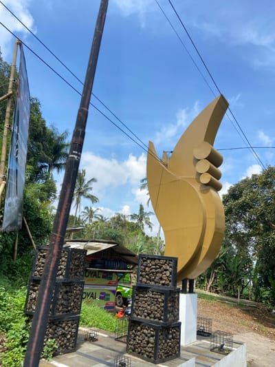 The image shows a large, golden, abstract sculpture that resembles a stylized flame or feather. The sculpture is mounted on a white concrete base with two black metal cages on either side. Behind the sculpture is a small, open-air shop with a green truck and a sign that reads "ROYAL." The shop is constructed of wood and has stacked stone walls. The sculpture is located in the rural, forested area of Cisarua, Indonesia, at latitude -6.839 and longitude 107.552. The sculpture is made of metal and is painted gold. It appears to be a public art installation or a decorative monument. The image was captured during daylight hours, under a clear blue sky with scattered white clouds. The ground is covered in grass and dirt. There are no people in the image.