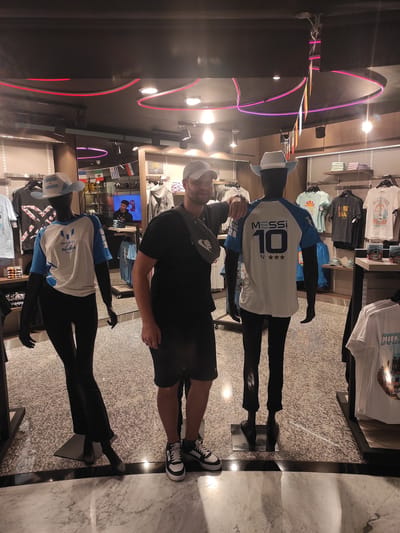 A man in a gray baseball cap, black t-shirt, and black shorts stands between two mannequins in a store, likely a gift shop at Aeropuerto Internacional Ezeiza, Argentina. The man has his right arm resting on the shoulder of the mannequin to his left. He is wearing black and white sneakers. The mannequin to his left is wearing a white and light blue shirt with "MESSI" and "Hard Rock" printed on it, and a white hat with light blue accents. The mannequin to his right is wearing a white shirt with "MESSI 10" and three stars printed on the back, and a white hat. The store has racks of clothing and display shelves. Flags are visible hanging from the ceiling. The floor is tiled with a speckled pattern.