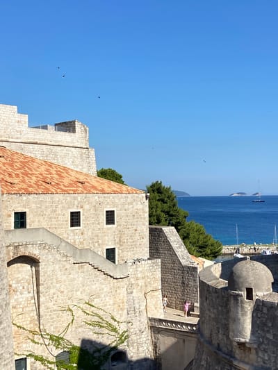 The image shows a view of the walls of Dubrovnik, Croatia, on a clear, sunny day. The foreground is dominated by the stone fortifications of the Old Town, featuring weathered stone blocks, arched doorways, and a tiled roof on one of the buildings. A staircase winds upwards along the wall, and some greenery is climbing up the stonework. Two women are visible walking on a walkway along the top of the fortifications. In the background, the deep blue Adriatic Sea stretches out, dotted with a few islands and a sailboat. The sky is a bright, cloudless blue with a few birds in flight. The scene evokes a sense of history and a Mediterranean coastal atmosphere.