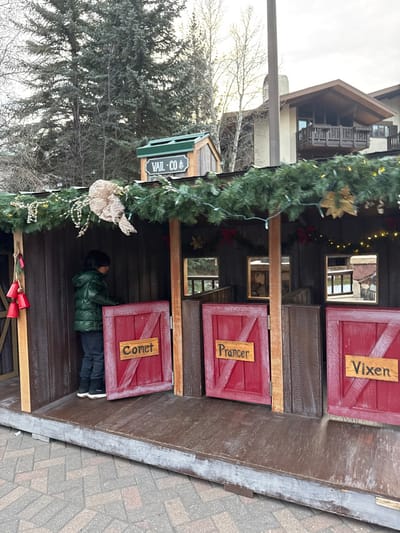 A child in a green jacket peers into a stall labeled "Comet" on a festive wooden structure. Above, doors are labeled "Prancer" and "Vixen". Garland with lights and a large bird adorns the side of the structure. A "Vail Co" sign hangs nearby. The structure has decorative mirrors above the doors. The backdrop features buildings and bare trees on an overcast day. The ground is paved with patterned bricks.
