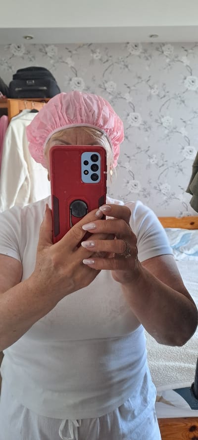 The image shows a person taking a selfie in a bedroom. The person is wearing a pink shower cap and a white t-shirt. They are holding a red smartphone in front of them. The background shows a floral wallpaper and a bed. The scene appears to be indoors, possibly in a home. There are no specific indicators of the city of Castlebar, Ireland.