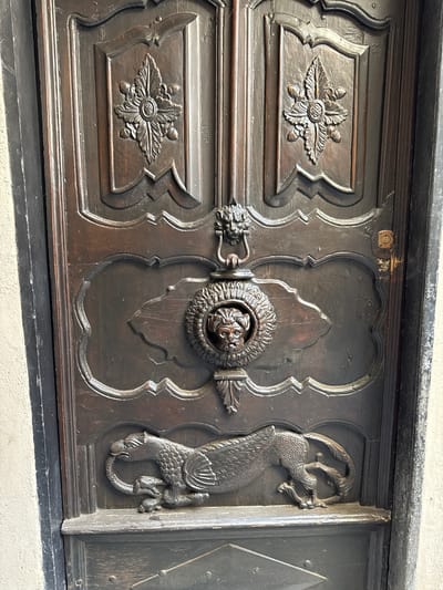 A close-up, eye-level, vertical shot shows the highly ornamented wooden double doors of a building in Apricale, Italy. The dark wood doors have elaborate carved details, including floral motifs, a lion's head knocker, and a mythical creature at the bottom. The knocker is centrally located, featuring a ring held by the mouth of a lion's head, which is itself within a circular frame. Below this, a winged creature, possibly a griffin, is depicted biting its own tail. The doors are divided into panels, with a light-colored, textured wall visible to the left and a dark frame along the right edge. The lighting is even, suggesting an overcast day or shade.