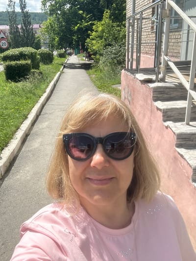 In Uchaly, Russia, a woman with blonde hair and sunglasses takes a selfie on a sunny day. She is wearing a pink t-shirt adorned with rhinestone designs. Behind her, a sidewalk runs alongside a grassy area and manicured bushes. In the background, a brick building with stairs and a metal railing is visible. The scene appears to be in a residential area.