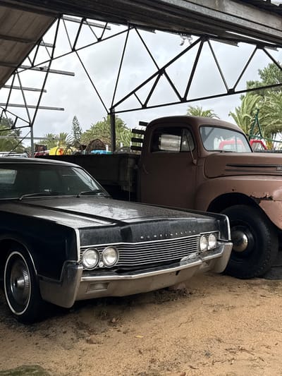 The image shows two vintage cars, a brown pickup truck and a dark-colored Continental car, parked close together in what appears to be an outdoor storage area in Kraaifontein, South Africa. The setting includes a metal structure possibly forming a shed or carport, and the ground consists of dirt and some vegetation. The sky is overcast, contributing to the overall dull lighting of the scene.