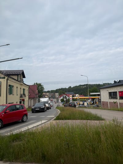 The image shows a street scene in Limanowa, Poland. In the foreground, tall grass is visible on the side of the road. A red hatchback car is driving on the left side of the road, passing a pedestrian crossing. Further ahead, a black Audi sedan is stopped in traffic, displaying Polish license plates KNS 822Z1. The road curves to the right, leading towards a Shell gas station with yellow and red signage. Several other vehicles are present, including a white truck. Buildings line the street, including a light yellow building with green shutters on the left and a building with a dark tiled roof and a prominent red sign on the right. The sky is overcast with scattered clouds. The overall scene depicts moderate traffic on a moderately developed urban street.