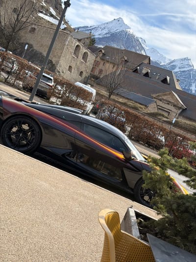 The image depicts a static urban and alpine landscape in Briançon, France, likely within or adjacent to the C Cité Vauban (Upper Town).

In the immediate foreground, a luxury sports car is parked on a light brown, granular paved surface. The vehicle exhibits a unique multi-chromatic paint finish, transitioning from dark brown/maroon to vibrant orange/gold hues, with black trim elements. It features black multi-spoke wheels with prominent brake calipers. To the lower right, a portion of a yellow-orange perforated metal chair and green coniferous shrubbery within a planter are visible.

The midground features a low, bare-branched hedge running horizontally, behind which a street is implied. Several vehicles are partially obscured by the hedge, including a silver SUV and a silver van or camper. A tall, dark utility pole stands vertically on the left side of the frame. Beyond the street and hedge, a sloped embankment with sparse, dry vegetation and leafless trees ascends towards historical stone architecture. Prominent structures include thick-walled stone buildings with arched window openings and dark-tiled roofs. One building to the right exhibits a distinct classical pediment above an entrance.

The background is dominated by majestic, snow-capped mountains under a clear blue sky with scattered white cirrus clouds, indicative of an alpine environment. No people are visible, and no specific actions or interactions are depicted.
