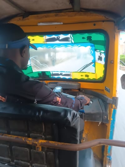 An eye-level, indoor shot captures a person driving a yellow three-wheeled vehicle. The driver, wearing a dark jacket and black baseball cap with a blue brim, grips the handlebars. The interior of the vehicle is visible, including the black seat and a rusty yellow bar. The windshield is cracked. Colorful decorations adorn the window, with birds painted in black and blue waves above a white pane. Outside, there are glimpses of a building and a road.