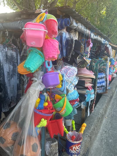 This image depicts a street-side stall in Bira, Indonesia, displaying an assortment of beach toys and apparel. The stall is set up along a concrete curb, with a corrugated metal roof providing shelter. Prominently featured are colorful plastic beach buckets and shovels, packaged in netting, in shades of pink, green, orange, and purple. Several hats, including straw hats and caps, are also visible, along with various clothing items such as patterned shirts and what appear to be sarongs or wrap skirts. Some larger items, possibly inflatable toys, are bagged in clear plastic. The stall is organized on racks and suspended from poles, suggesting a temporary or mobile setup common in bustling tourist or market areas. The environment appears to be outdoors, under the shade of trees, with dappled sunlight filtering through.