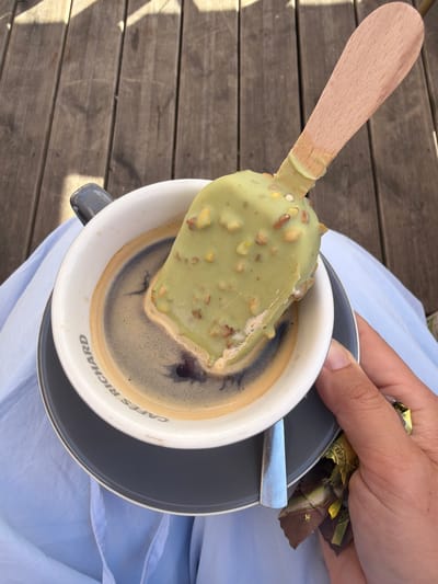 In Vernon, France, on a wooden deck, an ice cream bar with a green coating and nuts is submerged in a cup of black coffee. The cup is on a saucer, and a person's hand is holding the saucer and some wrappers. The scene suggests a moment of leisure and indulgence, possibly at an outdoor cafe.