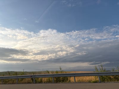 The image depicts a rural landscape with a vast sky filled with dynamic clouds. In the foreground, a metal guardrail separates a paved surface, likely a road, from a grassy embankment and fields beyond. The fields appear to be agricultural, with patches of dry golden vegetation and greener areas visible. The sky transitions from a clear blue at the top to a more overcast and dramatic display of clouds in the middle and lower sections, with sunlight breaking through in some areas, creating a contrast of light and shadow. The scene is likely captured from a moving vehicle, given the perspective and slight motion blur. The location is identified as Rupkite, Bulgaria.