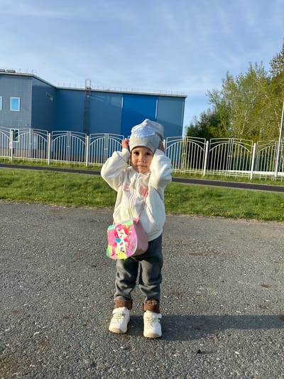 A young child stands on an asphalt surface near a grassy area bordered by a white metal fence. Behind the fence, a blue industrial building with multiple windows is visible. The child is wearing a gray hat with a sequined decoration, a white jacket, gray pants, and white shoes with brown accents. She carries a pink purse with a unicorn design. The sky is light blue. The location is in or near the city of Uray, Russia.