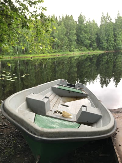 A high-angle, close-up, eye-level, vertical shot shows the interior of a grey and green plastic boat, with its exterior side resting on the ground. The boat is positioned diagonally on the right side of the frame, with its bow pointing upwards and towards the left. The boat’s interior is visible, featuring molded seats and compartments. A yellow scoop is nestled in one of the compartments. The boat's exterior is a dull grey on the inside and a vibrant green on the outside.
The background is dominated by a reflection of a dense forest with tall trees in a body of water. The water's surface is calm, creating a mirror-like effect of the trees and the sky. The reflection shows a straight line in the distance, which might be a dock or a path. The lighting suggests it could be overcast or late afternoon. The scene is peaceful and appears to be in a natural, possibly rural, setting. There is no text visible in the image.