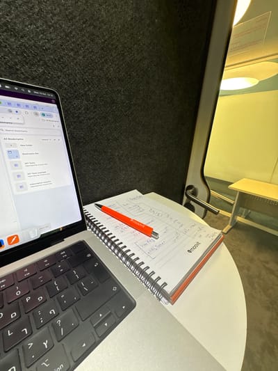 The image displays an indoor workspace in Nes Ziona, Israel. A silver MacBook Pro laptop occupies the left side of a light-colored, curved desk. Its screen shows a Chrome browser interface with "All Bookmarks" and the system time "Wed 15 Oct 13:10." The laptop's keyboard features both English and Hebrew characters. To the right of the laptop, a spiral-bound notebook with white lined paper and an orange spine is open, displaying handwritten notes, a grid, and a "moovit" logo on the lower right of the page. An orange "moovit" branded ballpoint pen rests diagonally across the open notebook. The background immediately behind the laptop and notebook is a dark grey, textured acoustic panel. To the right, a light-colored door frame with a metallic handle is visible, leading into an adjacent area characterized by bright yellow lighting and a light-colored rectangular table. No individuals are present in the scene.