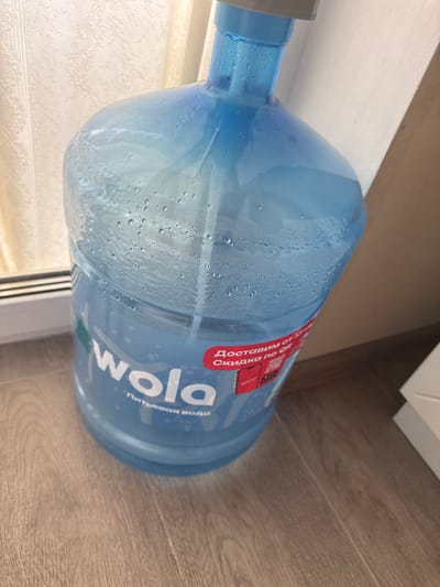 A large, blue plastic bottle of water is shown from a slightly elevated angle, resting on a wooden floor. The bottle is condensation-covered, indicating it is cold. The label on the bottle is predominantly white with some red accents and text. The text "wola" is prominently displayed in large white letters. Below "wola", smaller text in Russian reads "питьевая вода" which translates to "drinking water". There is also a red circular graphic with some red text within it, likely a logo or promotional element. Part of the label shows a red graphic resembling a carton with a handle and a red circle. The background consists of a light-colored wall and a wooden floor. The scene appears to be indoors, likely a home or office setting, given the presence of the floor and wall. There are no people visible in the image. The lighting suggests it is daytime, but no specific time of day is indicated. The location is identified as Romashkovo, Russia.