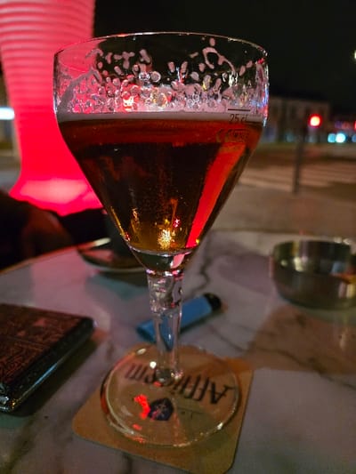 An eye-level shot captures a half-full glass of beer resting on a coaster, likely in a dimly lit bar or pub setting at night. The image is oriented vertically, but depicts a glass in a natural, slightly angled position as if on a table.

The main subject is a tulip-shaped beer glass containing a golden amber liquid with effervescence. A significant amount of white foam clings to the interior rim of the glass, creating an intricate pattern of bubbles. A measurement mark "25 cl" is clearly visible on the side of the glass, indicating a quarter-liter serving.

The glass sits on a round, light-brown coaster. Through the base of the glass and reflected on its surface, the brand name "AFFLIGEM" is distinctly legible, along with a visible coat of arms or logo, identifying the Belgian Abbey beer.

On the surface beside the glass, a small, bright blue lighter is partially visible. Further to the right, a dark, rectangular object with a patterned texture, possibly a wallet or a small book, lies on the table.

The background is dark and out of focus, featuring blurred points of colorful light, including reds, blues, and greens, contributing to a vibrant, nightlife atmosphere. The table surface appears light-colored, possibly marble or a similar material. The overall scene suggests a moment of relaxation and enjoyment of a drink in an evening social setting.