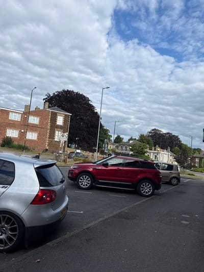 This image captures a street scene in Downham Market, King's Lynn and West Norfolk, United Kingdom, under an overcast sky with visible patches of blue. The perspective is from a low vantage point, looking across a paved street toward a row of buildings and parked vehicles.
In the immediate foreground, on the left, the rear three-quarter view of a silver Volkswagen Golf (Mk5 generation) is prominent. It features multi-spoke alloy wheels (BBS-style) and a partially visible UK license plate commencing with "JEZ". This vehicle is parked parallel to a raised curb.
Mid-ground, a red Range Rover Evoque SUV is parked on the asphalt road, positioned to the right of the silver Golf. The passenger side of the Evoque is visible. Further into the mid-ground, to the right of the Evoque, a light brown/beige small hatchback car is also parked.
In the background, to the left, a two-story brick building with several white-framed windows is visible. The right section of this building features darker, possibly stone, masonry with a distinct architectural style. Directly in front of this darker section, a stone monument supports a traditional town sign prominently displaying "DOWNHAM MARKET" in black text, beneath a heraldic crest and above a red banner depicting a figure. Behind this sign and building, a very large, dense, dark-leafed tree, likely a Copper Beech, stands out.
Further to the right in the background, a series of older-style buildings with varied facades, some featuring arched windows, line the street. Abundant green foliage from numerous trees and shrubs frames the scene, particularly along the distant treeline and around the buildings. Multiple streetlights are visible throughout the environment. The sky is predominantly covered by grey stratocumulus clouds, with a clearer blue patch showing in the upper right. No people are present, and no active interactions are depicted; the scene primarily shows parked vehicles and architecture.
