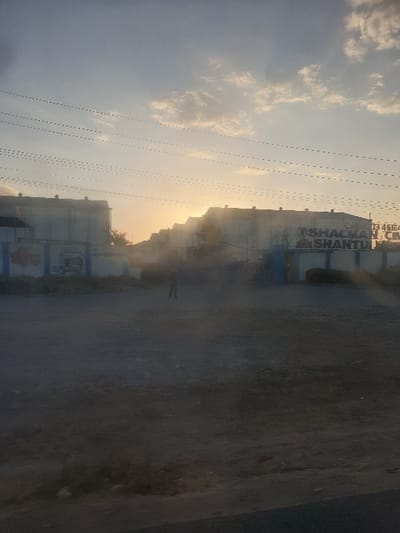 A hazy street scene with buildings lining the right side, partially obscured by dust or fog. The buildings display the text "SHACMAN SHANTU". A solitary figure stands in the middle ground. Overhead, a sky with scattered clouds and power lines creates a backdrop. The scene is bathed in a golden light, suggesting either sunrise or sunset. The overall tone is dusty and somewhat muted.