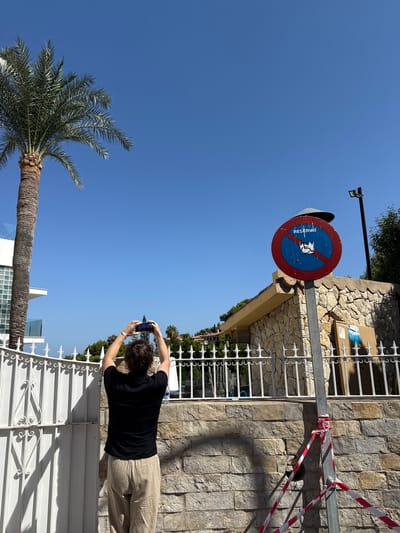 A person in a black t-shirt and tan pants stands with their back to the camera, holding a smartphone up to take a picture. They are positioned in front of a white metal fence, with a stone-clad building to their right. In the background, a palm tree stands tall against a clear blue sky. A "No Parking" sign, with the word "RESERVAT" visible, is mounted on a pole to the left of the building, partially obscured by a metal structure. Red and white striped tape is tied around a metal pole above the sign, suggesting some sort of construction or restricted area. The scene appears to be outdoors on a sunny day, likely in the afternoon, given the bright sunlight and clear sky. The location cues suggest Badia De Palma, Spain.
