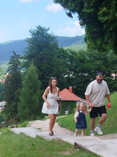 In Bușteni, Romania, a family of three walks down a paved path on a sunny day. A woman in a white polka-dot dress walks ahead of a man and a young girl. The man, wearing sunglasses and a light-colored shirt and shorts, holds the girl's hand. The girl, with blonde pigtails, wears a denim dress. They are surrounded by lush green trees and a distant mountain range. A building with a red roof is visible in the background.