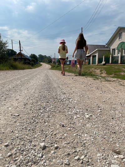 Two young females walk away from the camera down a gravel road in the rural village of Baldeyka, Russia. The female in the foreground, wearing denim shorts and a dark top, carries a pink bag with what appears to be flowers. The second female, slightly ahead and to the left, wears a yellow t-shirt and a pink hat. The road is lined with houses, trees, and utility poles. The sky is partly cloudy.