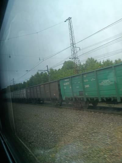The image depicts a train of hopper cars traveling on a railway track. The train is composed of several cars, some of which are dark gray, and others are bright green. The green cars have visible text and numbers printed on their sides. The railway track is covered with gravel. In the background, there is a dense line of green trees. Overhead, there are power lines supported by tall metal towers. The sky is overcast and appears to be a pale blue or gray. The image seems to have been taken from a moving vehicle, possibly another train, as indicated by the motion blur. The location is identified as Трансмаш Снт, Russia.