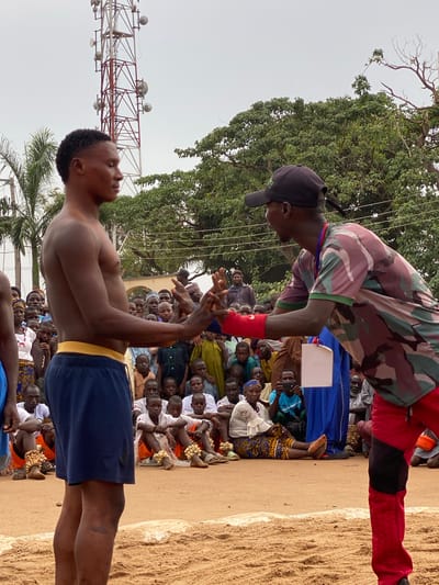 In an outdoor setting within Kafanchan A, Nigeria, two individuals are engaged in a hand-to-hand interaction against a backdrop of spectators seated or standing around a sandy area. One individual, shirtless and wearing blue shorts with a yellow waistband, stands facing the other. The second individual wears camouflage-patterned clothing on their upper body, red arm wraps, red trousers, and a black baseball cap. The two appear to be interlocking hands or wrists. Behind the subjects, a crowd of people of varying ages is visible. A tall communications tower is present in the background. To the left, there is a palm tree, and above, a large tree provides shade.