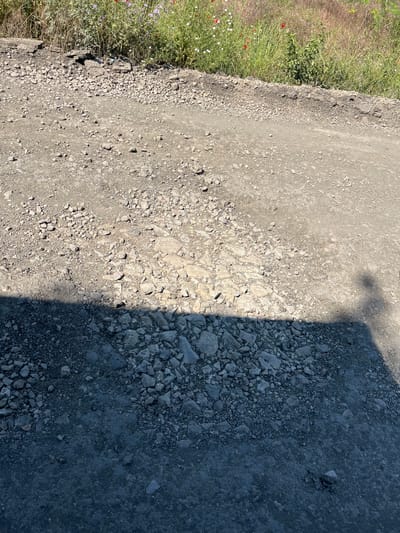 The image shows a rough, unpaved road or path in the city of Poroy, Bulgaria. The ground is covered with gravel and stones of various sizes. In the upper part of the image, there is a patch of dry grass and wildflowers, including some red poppies and white flowers, growing along the edge of the path. The lower part of the image is cast in shadow, revealing a darker, more compacted surface with scattered stones. The shadow suggests the presence of an object or person outside the frame, casting a silhouette of a head and shoulders. The overall scene depicts an outdoor, somewhat unkempt environment.