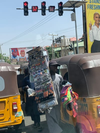 The scene depicts a bustling street environment in Hotoro, Nigeria, under bright daylight. In the foreground, a male street vendor, dressed in a grey long-sleeved tunic and black baseball cap, is positioned between two three-wheeled auto-rickshaws. He carries a large, rectangular display board on his left shoulder, laden with numerous small merchandise items including rings, earrings, keychains, and small leather pouches, individually packaged or displayed. In his left arm, he holds a bundle of colorful folded fabrics or scarves, while his right hand clutches a clear plastic bag containing light-colored, possibly wooden, objects.
To the vendor's left, the rear of a yellow auto-rickshaw with a Kano state license plate "KMC-521" is visible. To his right, a brown auto-rickshaw's side is present. Overhead, a set of traffic lights displays red arrow signals for left turns, accompanied by digital countdown timers showing "52" seconds. Power lines crisscross the upper portion of the image.
In the background, two large billboards are visible. The left billboard is political, featuring two male figures identified as Bola Ahmed Tinubu and Kashim Shettima, with the slogan "RENEWED HOPE...STRONGER NIGERIA." The right billboard, predominantly yellow, features a light-skinned female model with blonde hair, gesturing with her right hand, advertising a product related to "STAINS FRESHNESS." Distant buildings with red and green roofs are discernible under a clear sky.