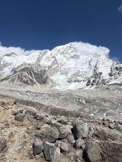 This is a vertically oriented outdoor photograph capturing a dramatic high-altitude mountain landscape in Khumjung, Nepal. The left side of the frame is dominated by a vast, deep blue sky, accented by small wisps of white clouds. A more substantial cloud formation hugs the upper slopes of the central mountain, suggesting active weather around the peaks.
The primary subject is a majestic, snow-capped mountain peak rising prominently. Its rugged surfaces are extensively covered in brilliant white snow and intricate glacial ice, exhibiting sharp ridges, deep crevasses, and textured icy slopes. The bright sunlight creates strong contrasts, illuminating the snow while casting deep shadows that emphasize the mountain's formidable topography.
To the right of the snowy peak, and extending into the foreground and midground, is a steep, barren rocky slope. This area features a jumble of large, dark grey boulders, smaller angular rocks, and loose scree. Traces of snow or ice are visible intermittently among the rocks, particularly in shaded crevices. The terrain is typical of a high-altitude, alpine environment above the treeline.
The scene depicts a bright, clear day, characteristic of good mountain weather, with the vibrant blue sky and strong illumination. There are no people, animals, or visible text in the image. The photograph conveys a sense of immense scale, natural grandeur, and the remote, raw beauty of the Himalayan wilderness.