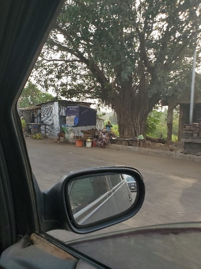 A view from inside a car features a side mirror reflecting a road with a double yellow line. Outside the car, a large tree dominates the scene, casting shade over a small roadside market stall. The stall is cluttered with various items and some orange buckets. The sky is overcast, and the image appears slightly blurry.